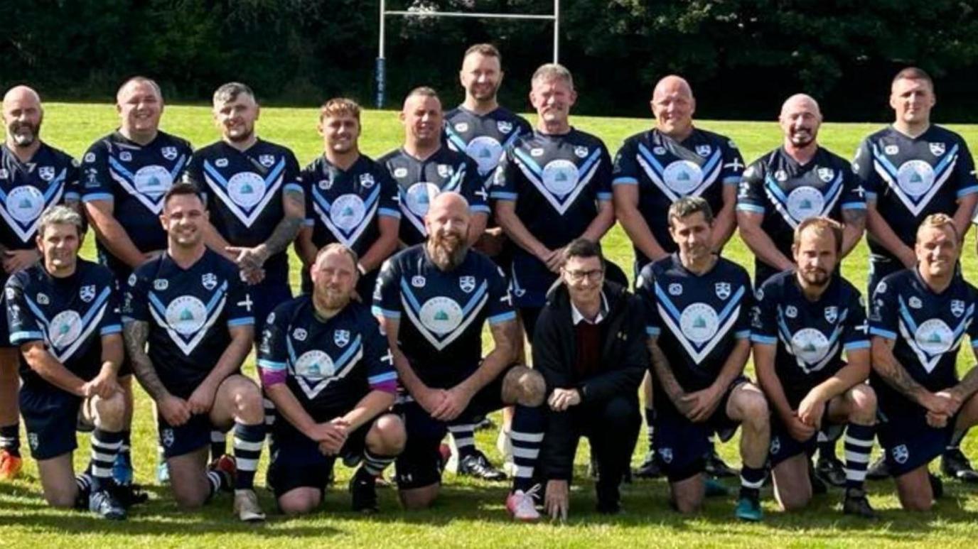 A team photo of the rugby club's players lined up in two rows on the pitch with rugby posts behind them. The first row is kneeling and the second row standing and the players are all clasping their hands together in front of them.