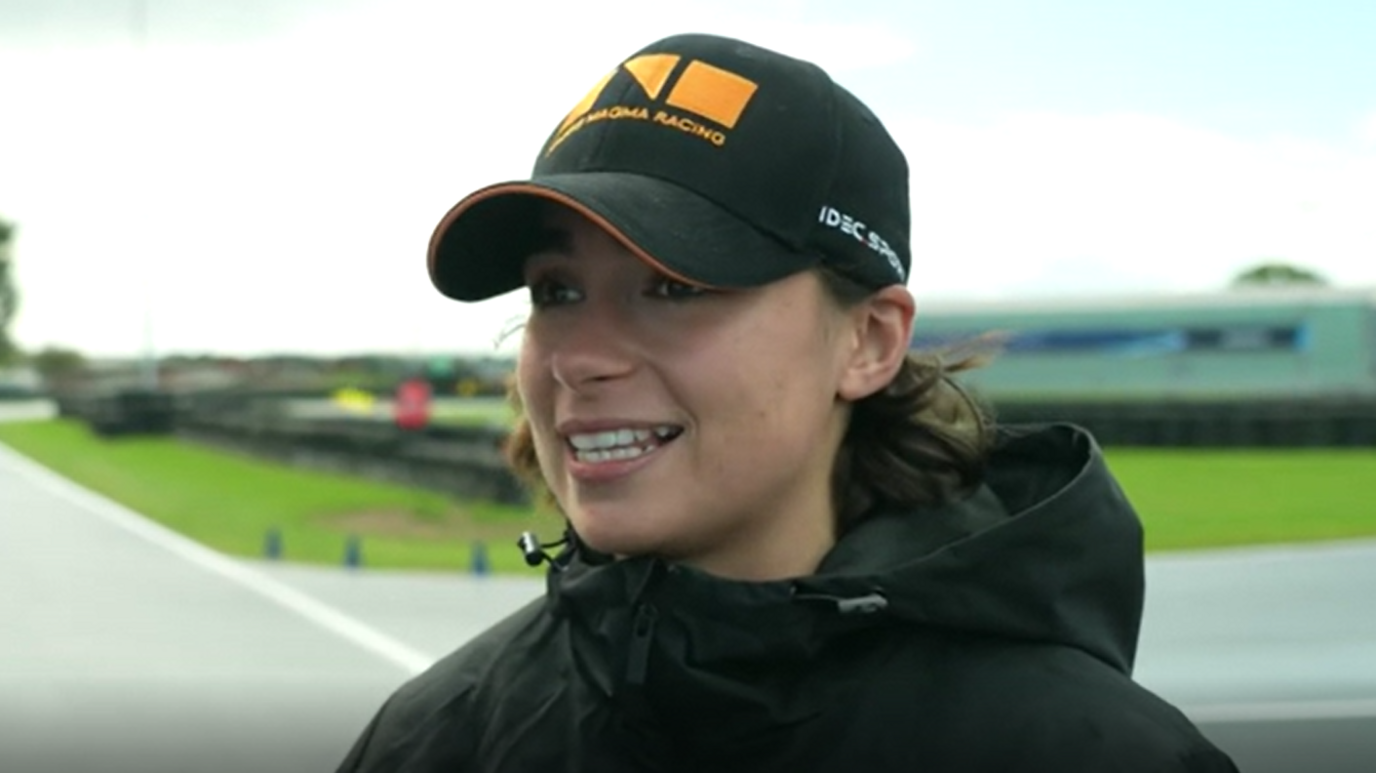 Racing driver Jamie Chadwick who is standing outside at the track at Silverstone. She is wearing a black coat with a hood and a black baseball cap.