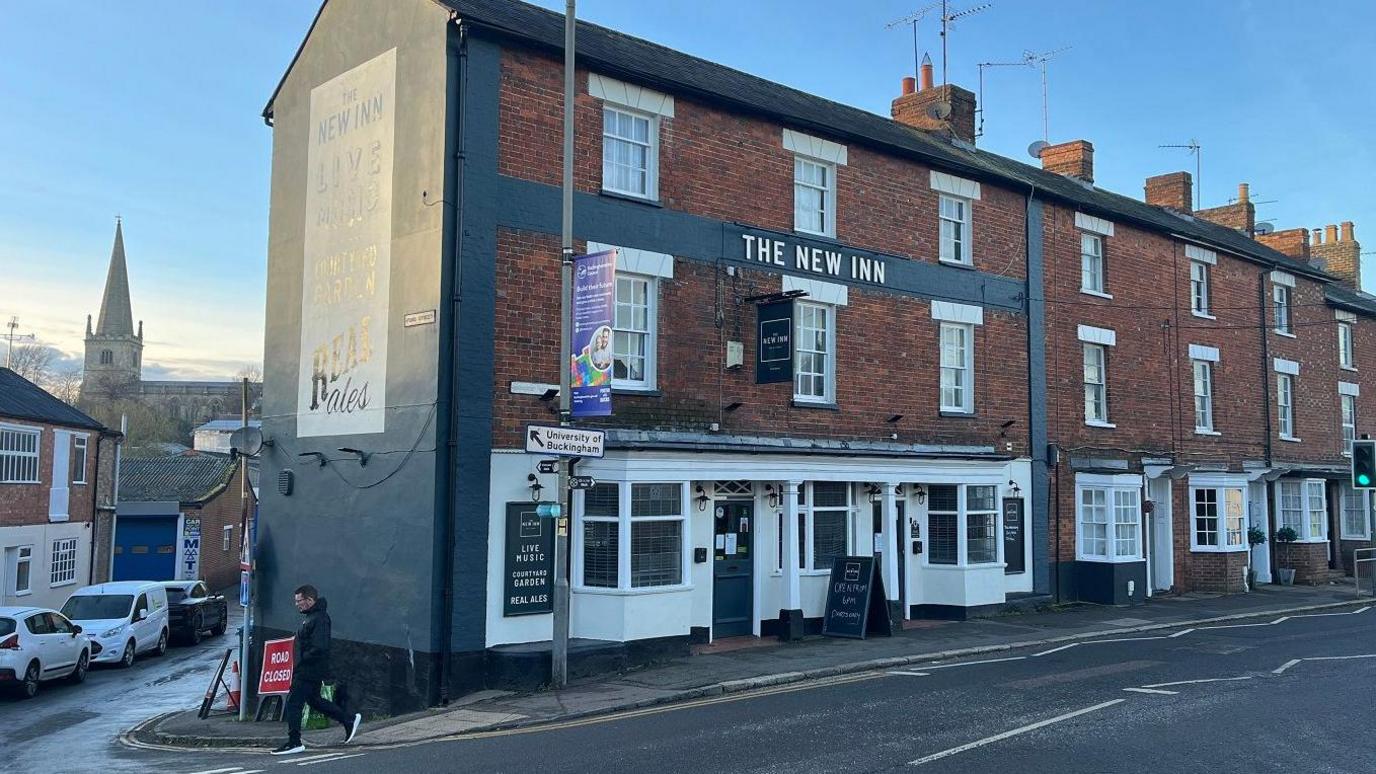 A street scene in Buckingham. There is a church in the background and the New Inn in the foreground. It is at the end of a row of traditional buildings  with red bricks and white sash windows.