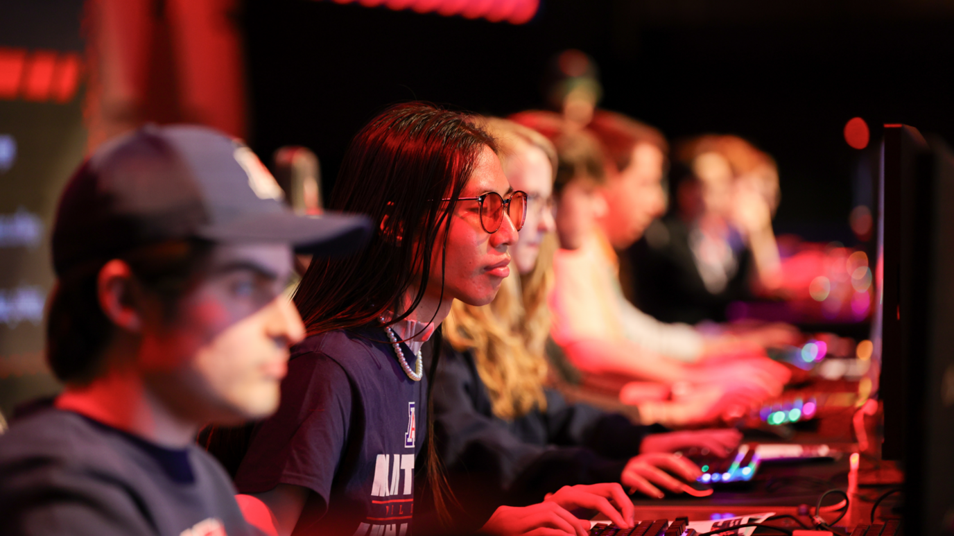 Players focused on their computers during a MEWC round in Las Vegas. The area is lit up with red light and only one person, along a line of people sat in front of computers, is in focus. They have long dark hair and are wearing tinted sunglasses.