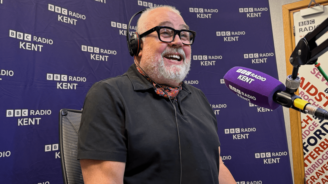 Cliff Parisi laughing into a purple microphone with "BBC Radio Kent" on the side. He is wearing a black polo, patterned cravat, glasses, and is in front of a purple backdrop with "BBC Radio Kent" written on it several times.