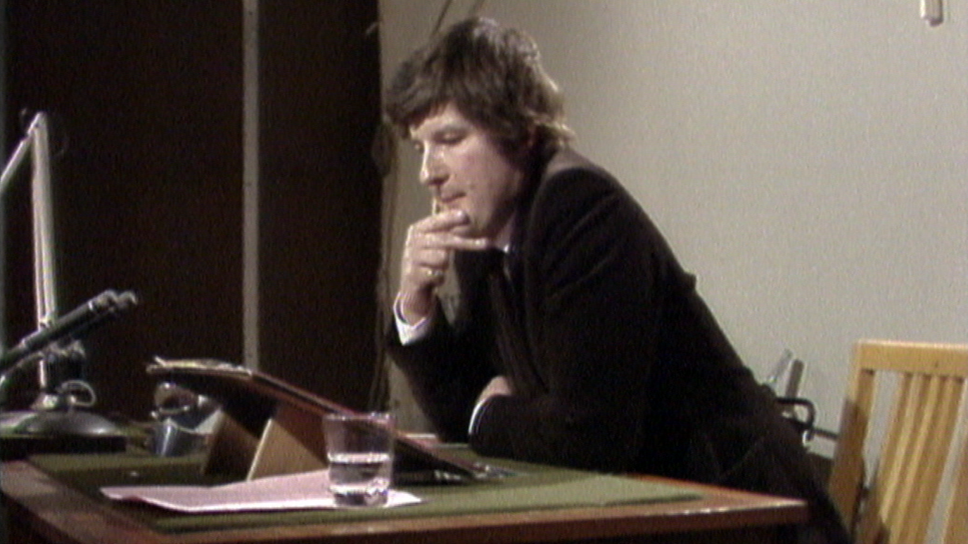 Reporter Kim Riley sitting at a desk, speaking live into a microphone. He is wearing a brown suit and white shirt.