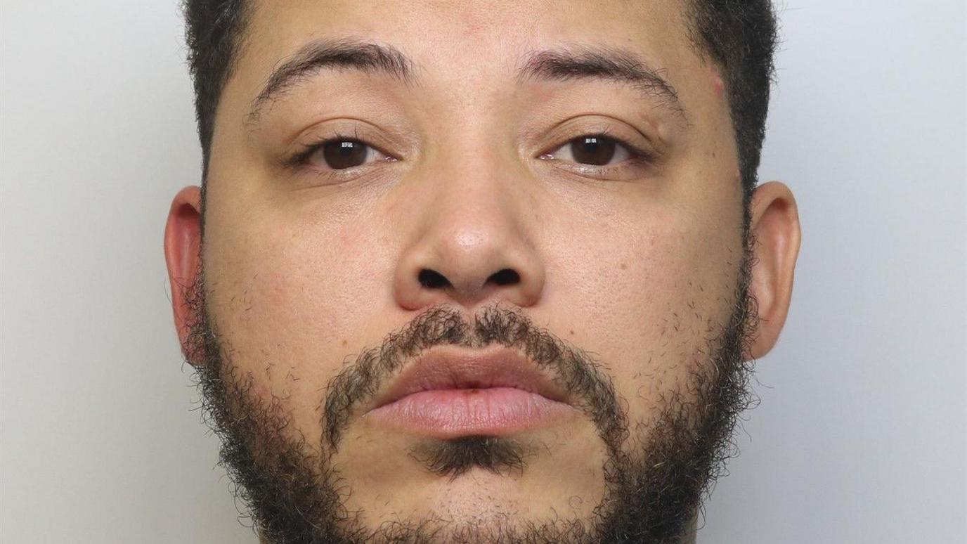 A man staring straight ahead with a blank expression. He has short, dark hair and a short beard. 