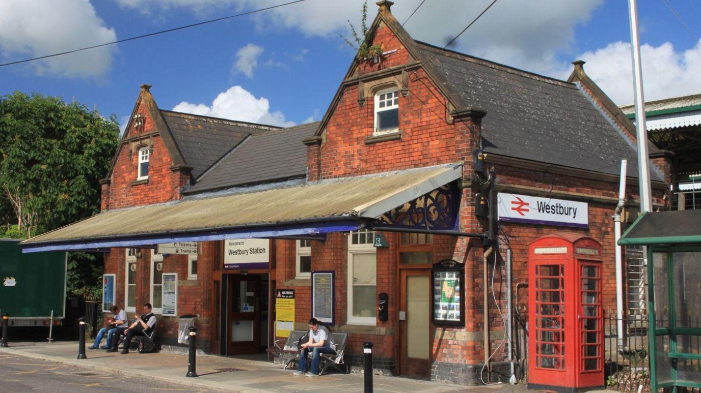 Westbury train station in Wiltshire. It is a small, old brick building with a plastic awning and a red telephone box outside the front. There are several people sitting on benches outside, waiting for their trains. The platform is not visible behind the building, but beside it are several trees.