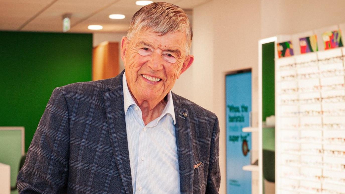 Man in a jacket which is blue and white and a light blue shirt. He has grey and white hair and is in a Specsavers store