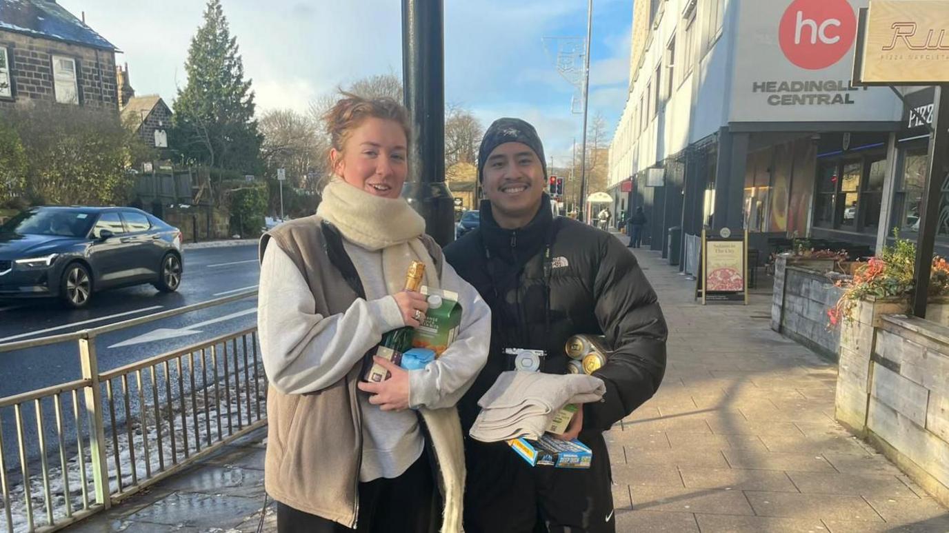 Rose Hubble and Lewis. They are both carrying shopping. Rose is wearing a cream scarf with a coffee-coloured gilet, while Lewis has a black padded jacket on with a black beanie hat.