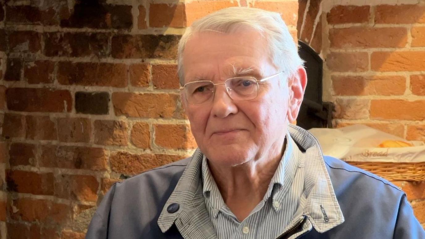 Richard Brown gives a TV interview indoors with an exposed brick wall behind him. He has white hair and glasses and wears a casual jacket over a striped shirt.