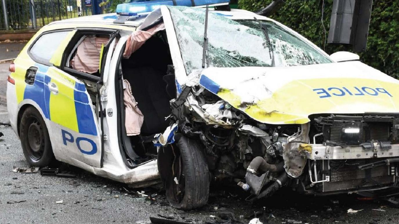 A very badly damaged police car, with a crumpled bonnet, smashed windscreen, and twisted body. The airbags have been deployed and there is a debris on the road