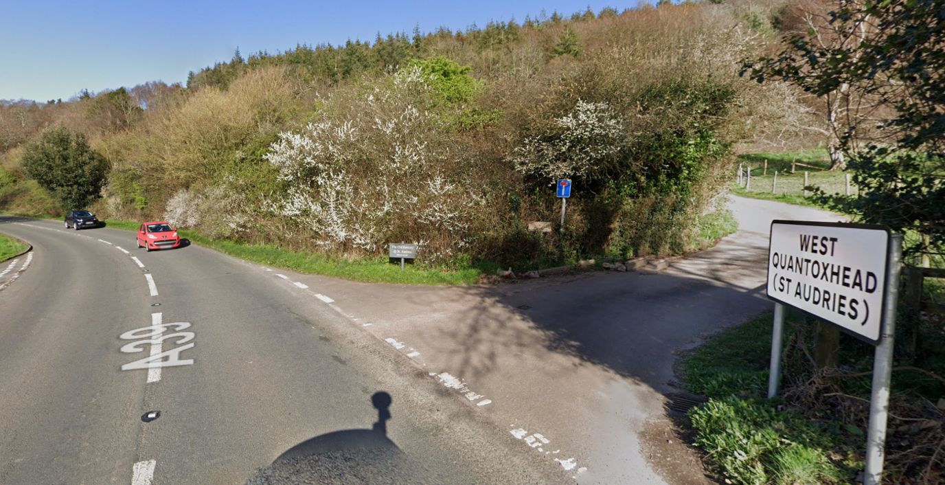 Google maps street view image of the A39 at West Quantoxhead. The A39 is winding round to the left and to the right a road veers up, with the West Quantoxhead sign at the bottom of the hill. 