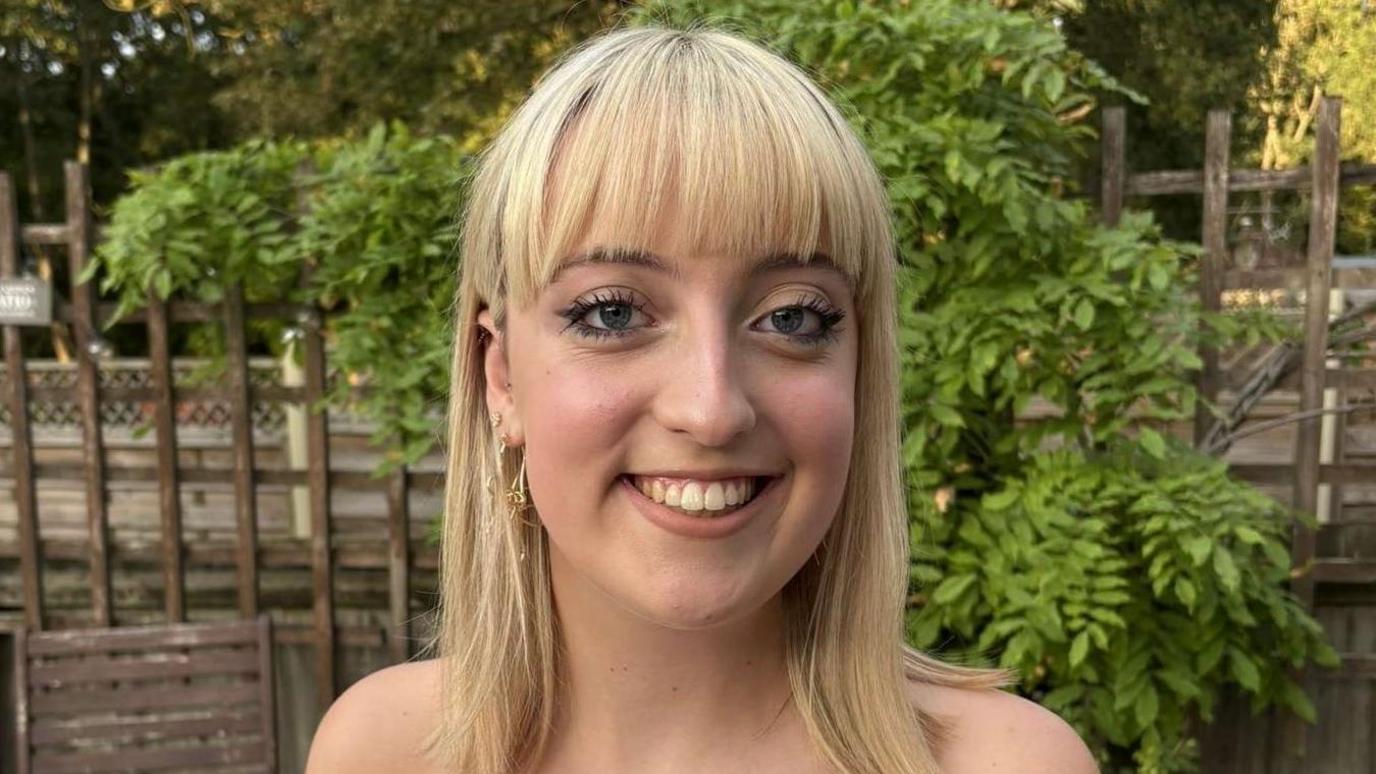 Imogen who has blonde mid length hair and a fringe looks at the camera smiling. Behind her is a brown garden fence and green leaves. 