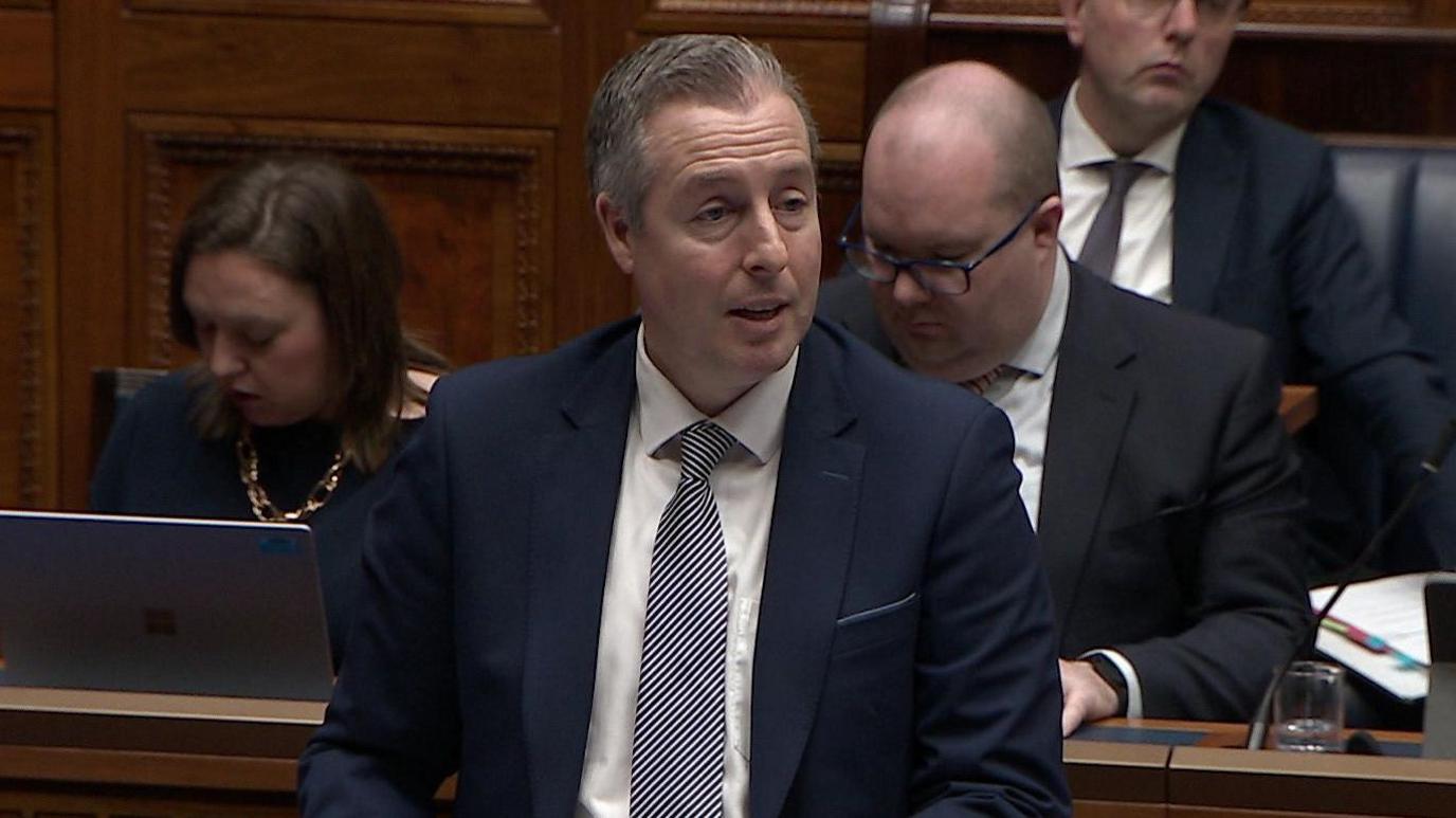 Paul Givan speaking in the Assembly. He has grey-ish hair, wearing a navy blazer, white shirt and strpid tie. Other MLAs are in the seats behind him.