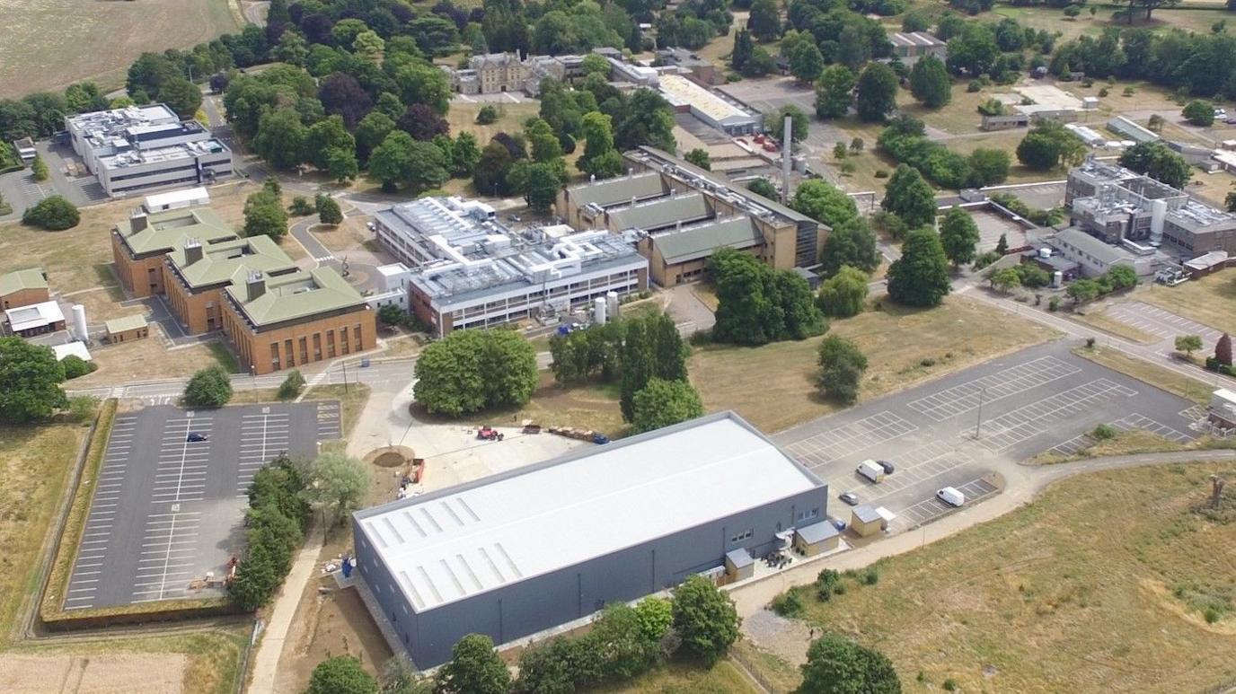 An aerial view of a large science park's buildings and car parks. It includes several large warehouses and factories. There are trees, shrubs and areas of grass between the buildings.