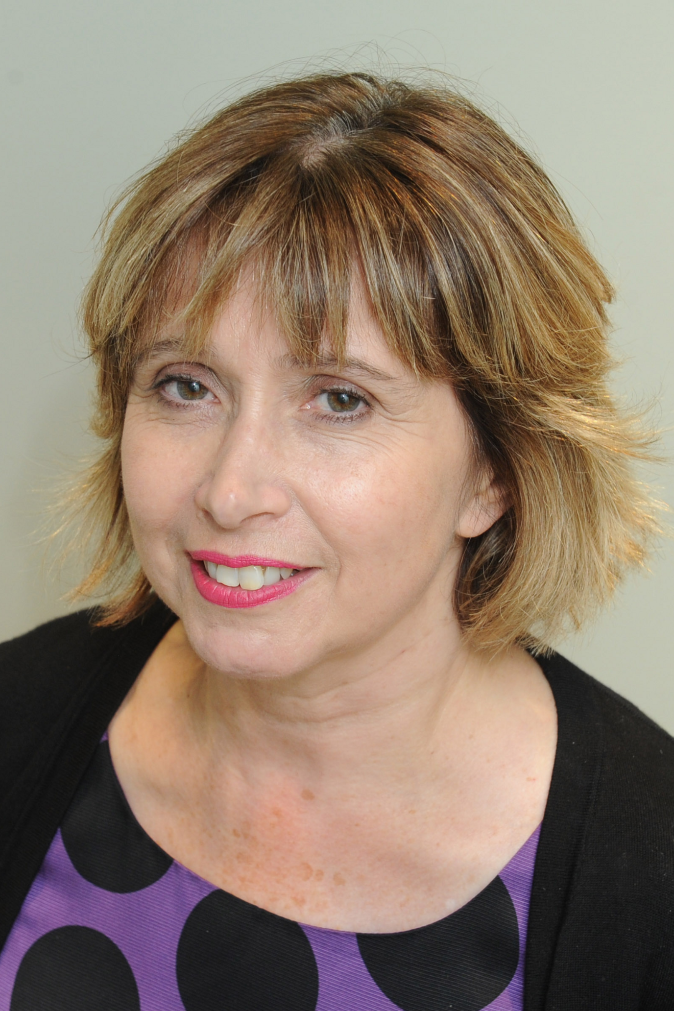 A woman with short blonde hair wearing a purple shirt with large black circles on it and a black cardigan smiles at the camera.