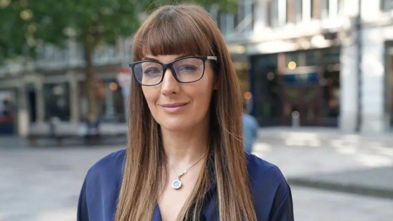 A woman with long brown hair and glasses stands on a Cardiff street, the background is blurred