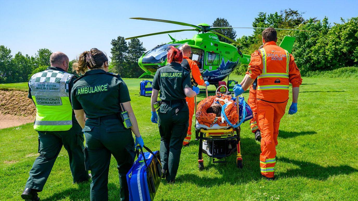 A group of five paramedics wearing green uniforms, orange jumpsuits with yellow hi-vis straps, and a yellow hi-vis vest. One is carrying a blue blag full of medical tools. They are conducting a simulation exercise, and are walking towards a green and blue helicopter which is parked on the grass of a golf course. The three paramedics in the front are wheeling along a gurney with a patient on it.