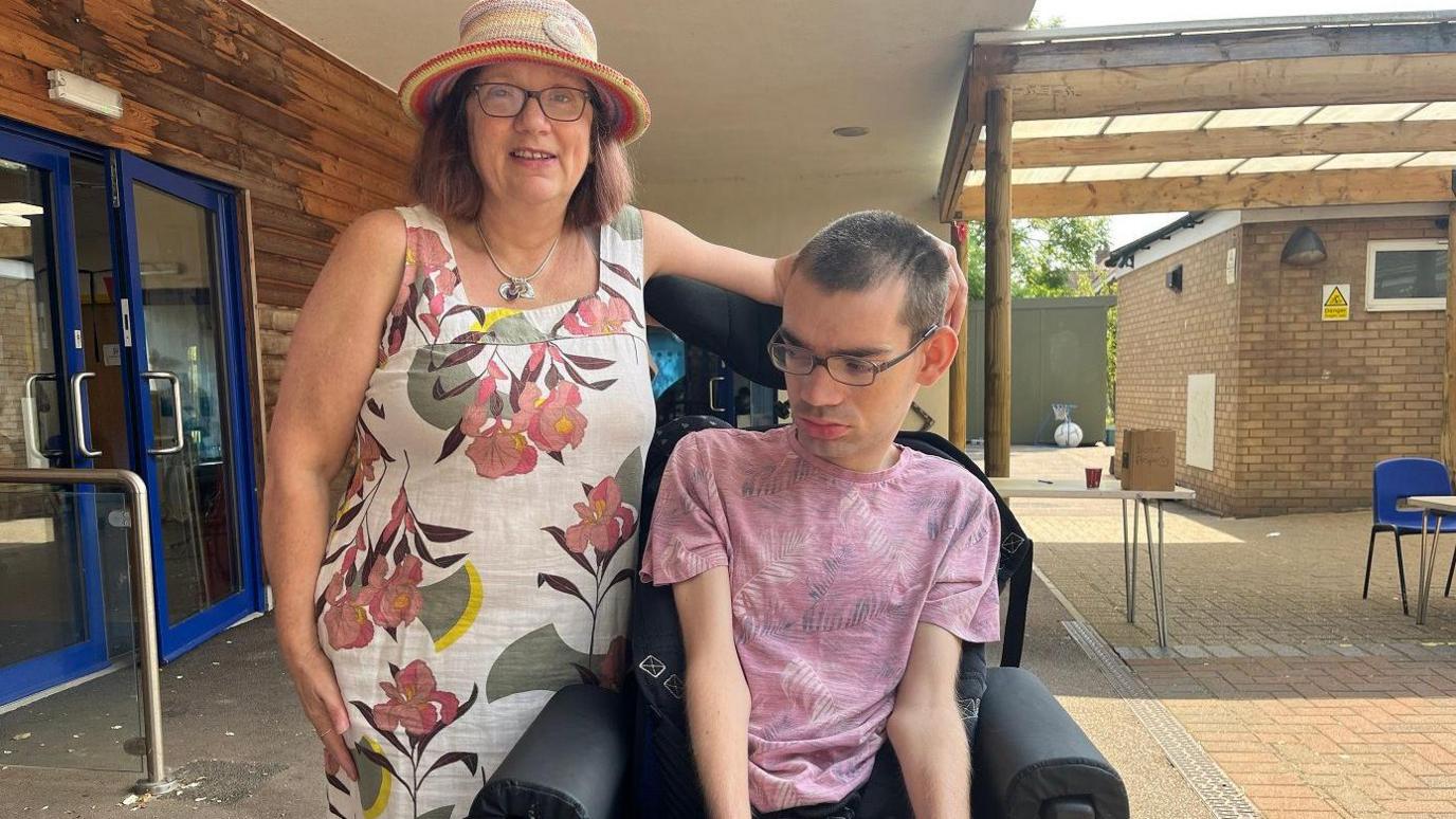 Lilian, wearing a hat and sundress, next to Jamie wearing a purple t-shirt sitting in his chair