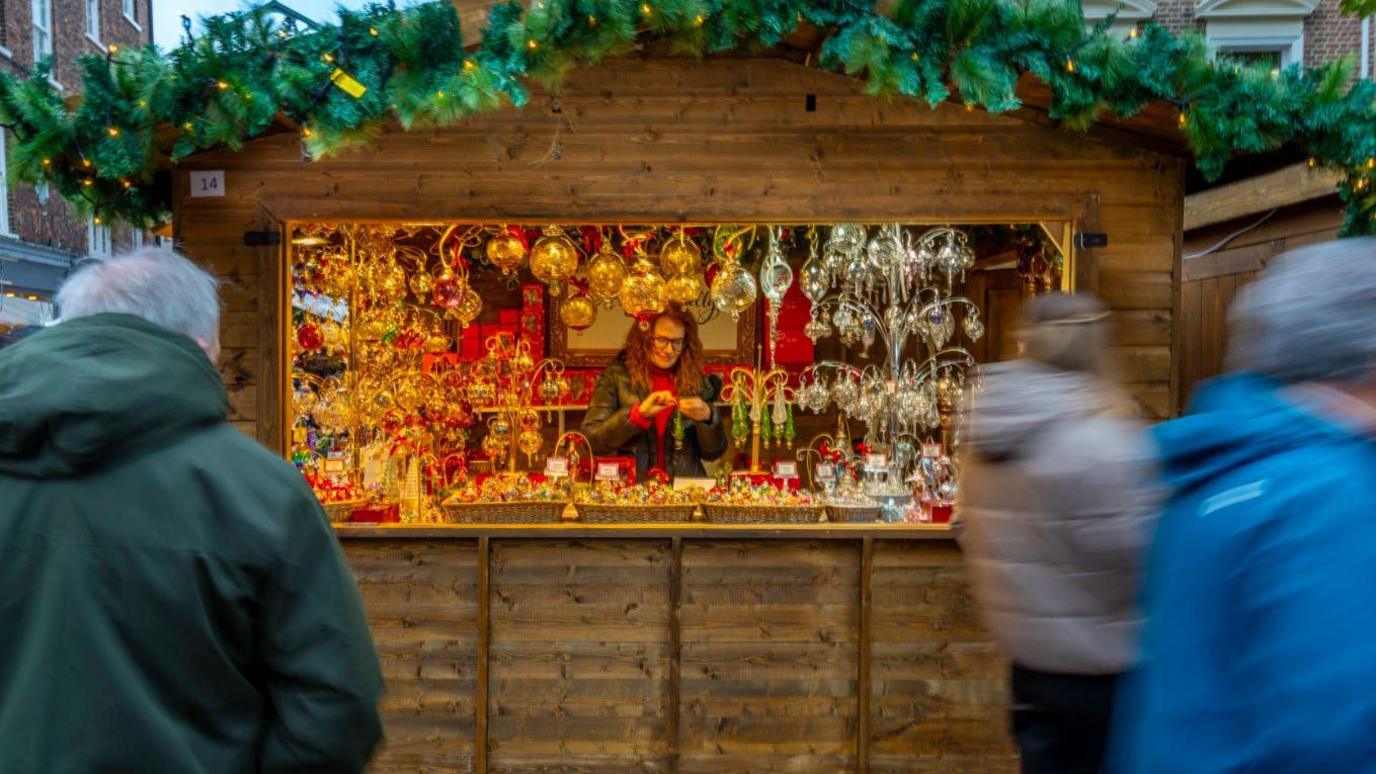 A wooden hut used as a market stall is filled with Christmas decorations. A woman with red hair and wearing a red sweater stands inside the kiosk. Blurred images of people walking past the attraction are in the foreground.