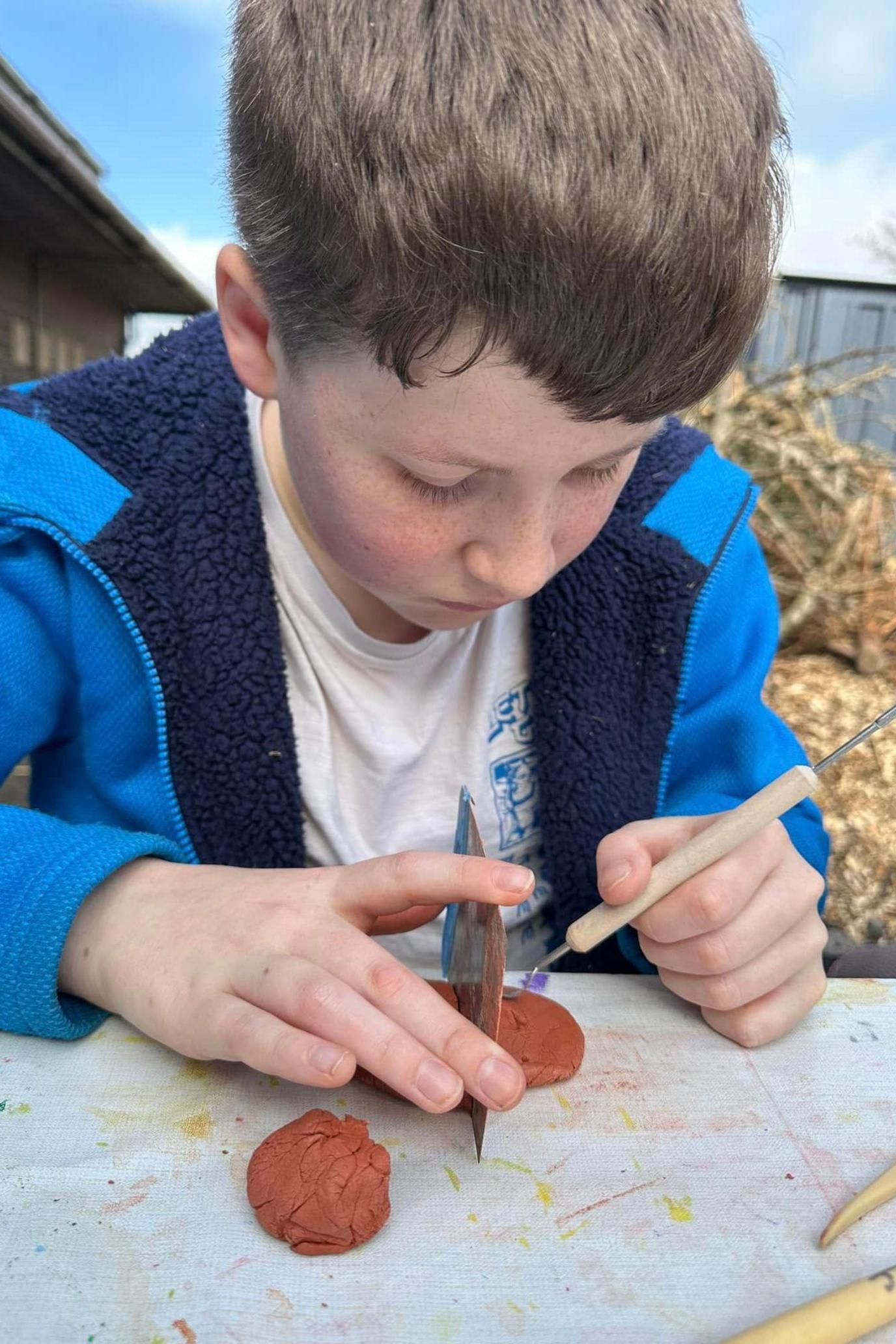 A young boy with brown hair sat at a table holding model clay. He had his head down as he works on the craft. He is wearing a white t shirt and blue hoodie and behind him are stacks of hay bails.