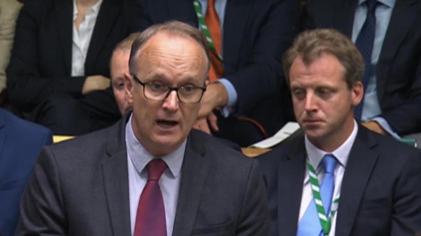 Dr Neil Hudson, the Conservative MP for Epping Forest, standing in the House of Commons addressing parliament. He has thinning hair, large rimmed glasses and is wearing a purple tie, light blue shirt and dark blue jacket.