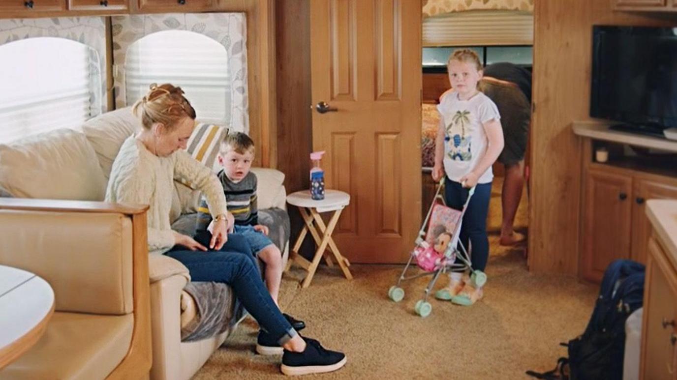 Rachael and her two kids in the living room of a trailer with yellow pine paneling a grey leather sofa and flatscreen TV