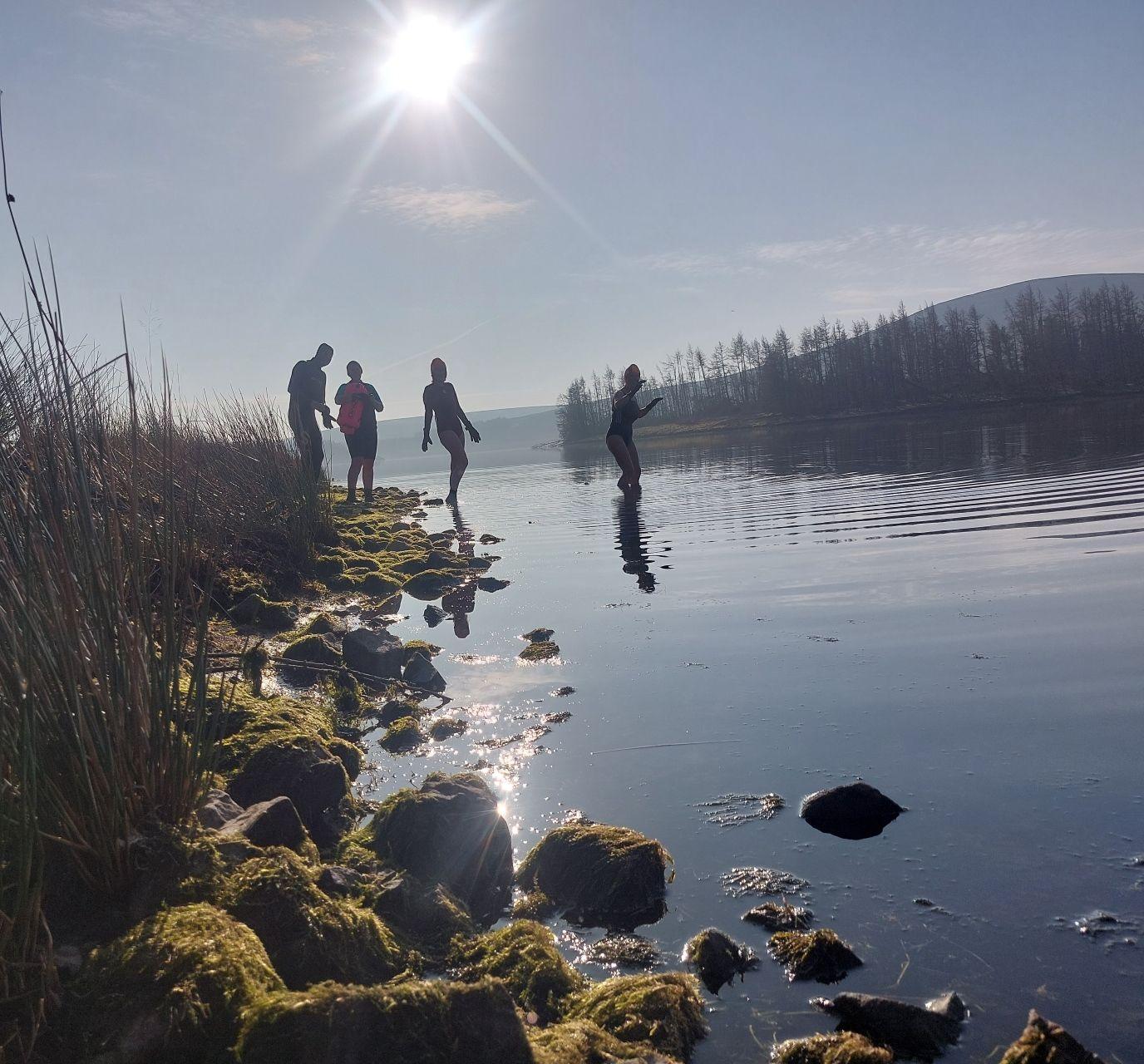 Four wild swimmers stand in a reservoir, with stones lit by the sun in the foreground.