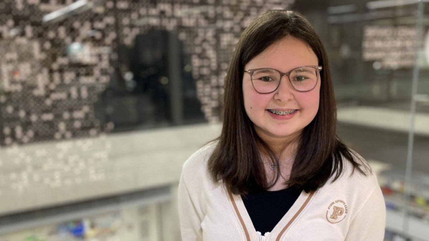 A teenage girl with dark hair smiles at the camera, wearing a white cardigan