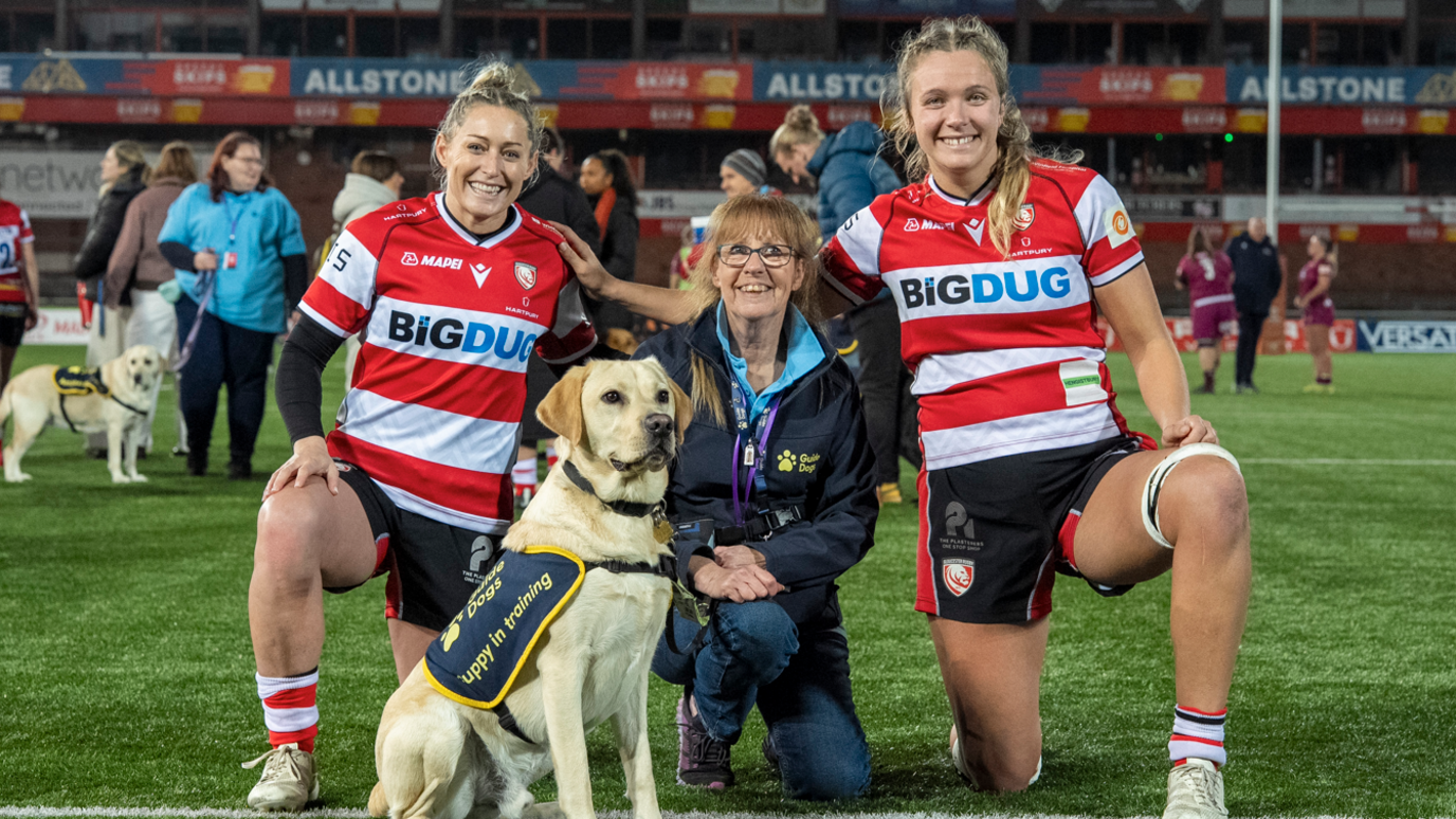 Image of the co-captains posing for a photo next to a volunteer and puppy. The co-captains are in their red and white jerseys and the volunteer in a navy blue jacket and jeans.