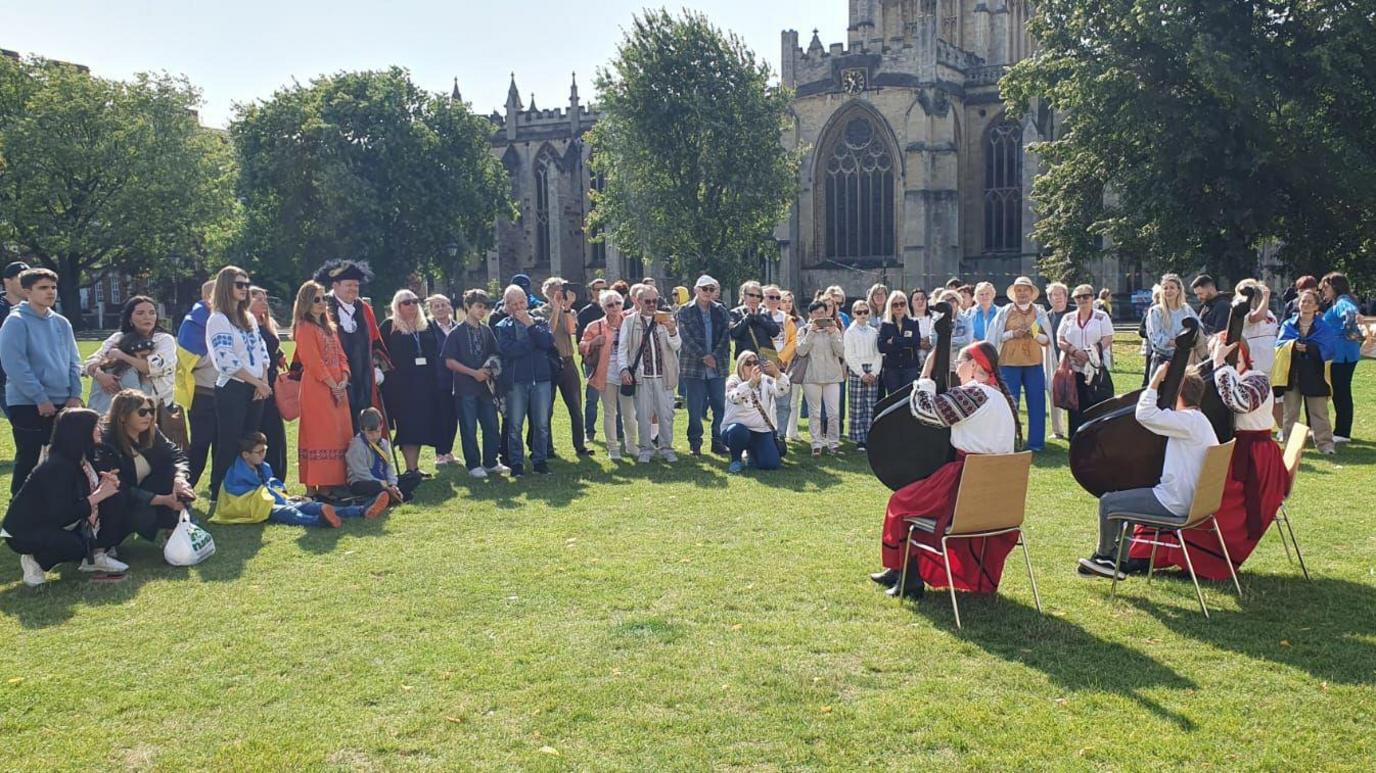 People wearing traditional outfits playing music to a line of people standing outside