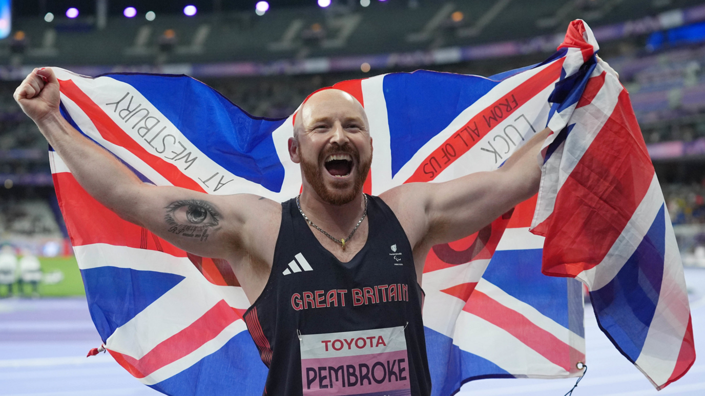 Dan Pembroke in his navy blue Great Britain singlet smiling and carrying a Great Britain flag at the Paralympics in Paris