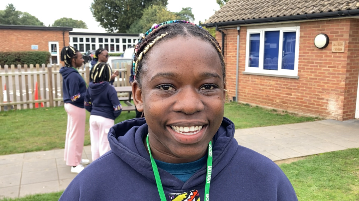 Elizabeth has colourful braids in her hair and is smiling into the camera. She is wearing a navy hoodie and has a green lanyard around her neck. In the background are other members of the choir, talking.