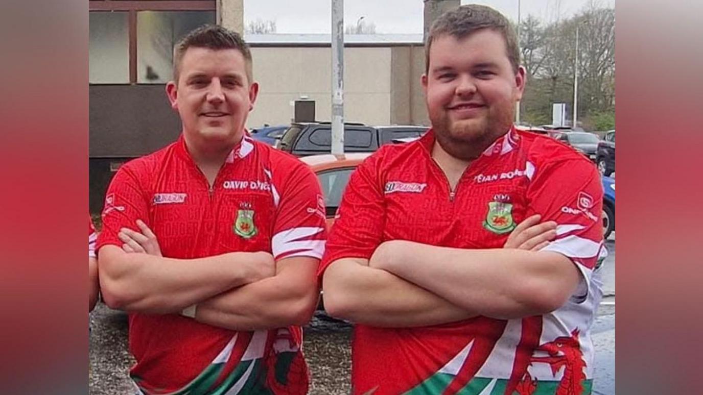 Dau ddyn yn sefyll gyda'u breichiau wedi croesi o'u blaenau nhw. Ar y chwith mae David Davies sydd gyda gwallt brown byr. Ar y dde mae Deian Roberts sydd gyda gwallt brown byr a barf brown. Mae'r ddau yn gwisgo crys-t coch gyda draig Cymru arno.