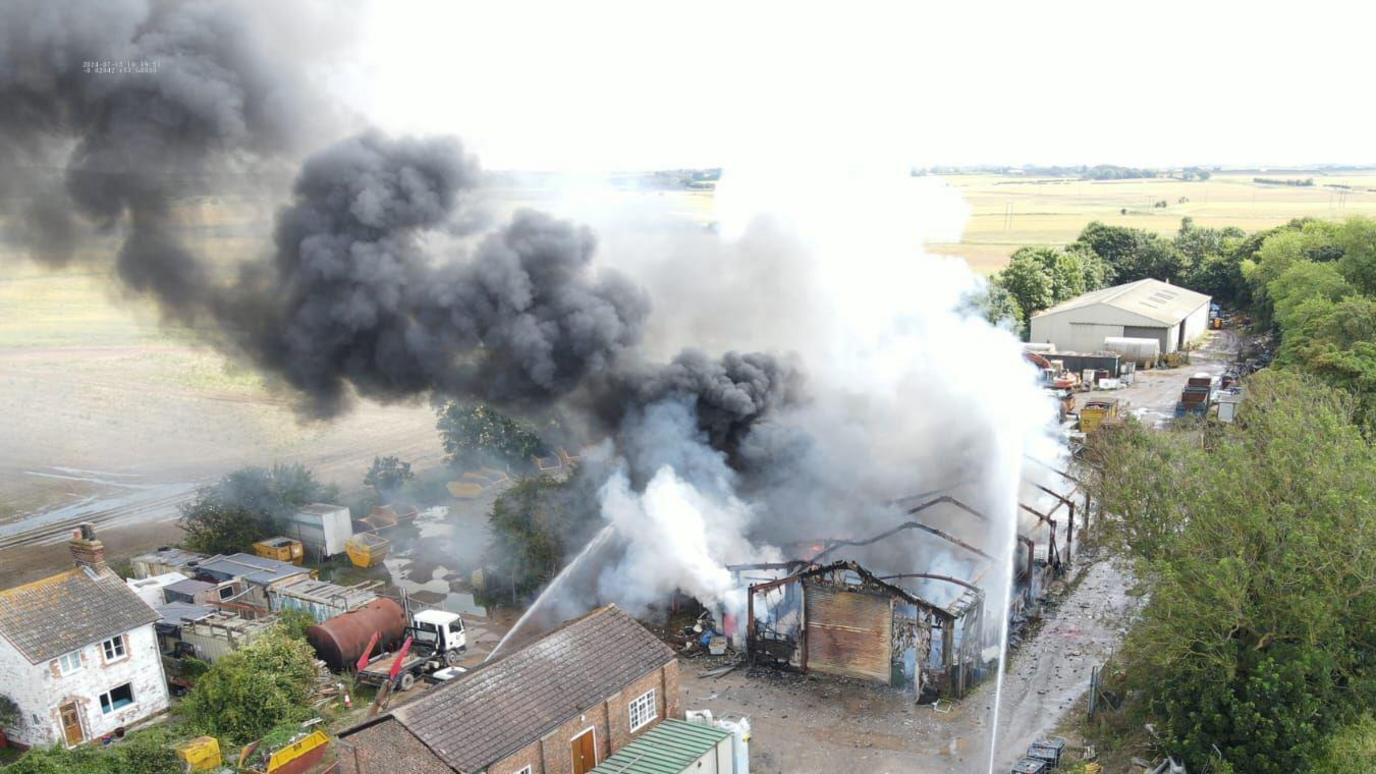 Smoke billowing into the air from a fire-damaged building at a commercial unit, which is surrounded by fields and is also close to a house and other buildings