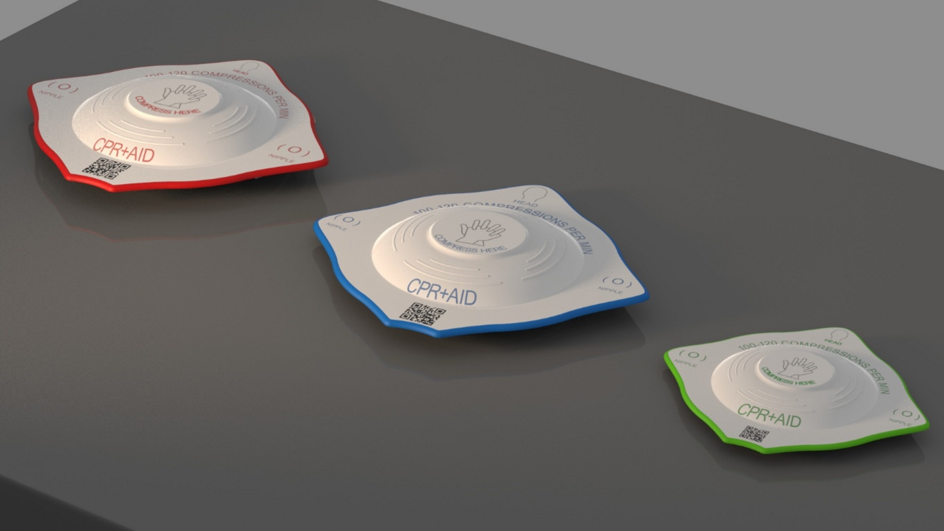 Three CPR+Aid devices, designed by Harry Wragg, each a different size and colour on a dark grey table.