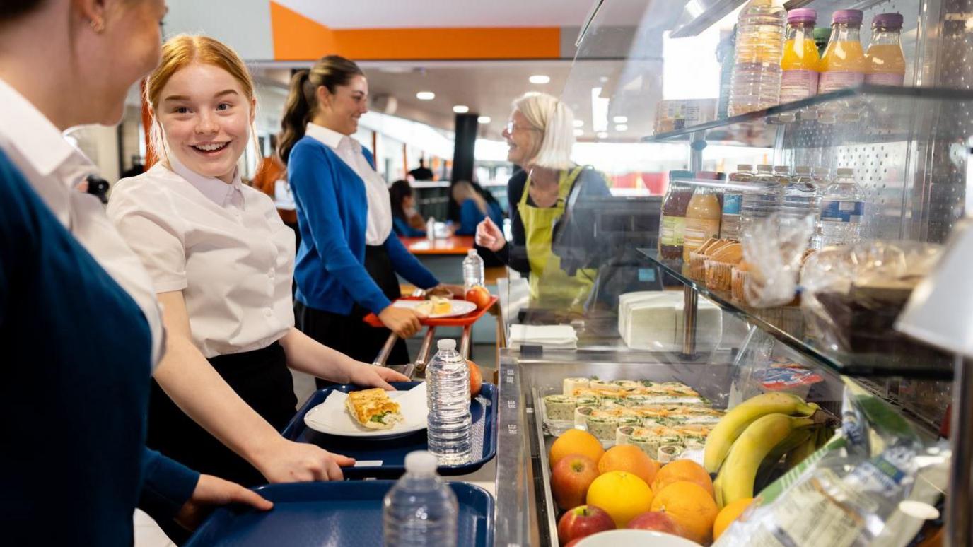 Three schoolgirls queuing to pay for their school meals. There are number of healthy options on glass shelves in front of them, including a selection of fruit.