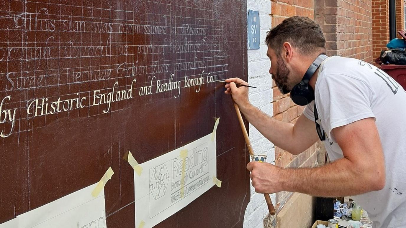 Man painting calligraphy on to a wall that has been painted dark red. The words read "...by Historic England and Reading Borough..."