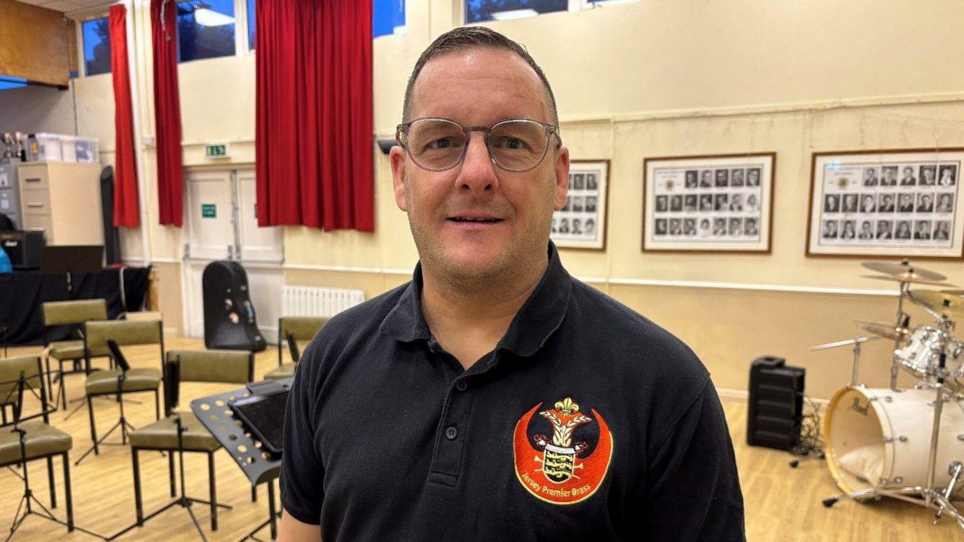 Jason Mildren is wearing a dark polo shirt and spectacles. He is looking at the camera and slightly smiling. He is standing in a room with chairs set out for the band, and music stands. There is a drum kit behind him near a wall.