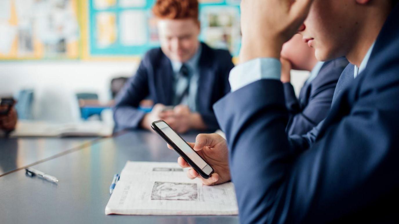 Boy using a smartphone in a classroom. Anonymised photo so you can only see his arms leaning on a desk and he's looking downwards at the phone screen. Behind him is another boy looking at his phone.