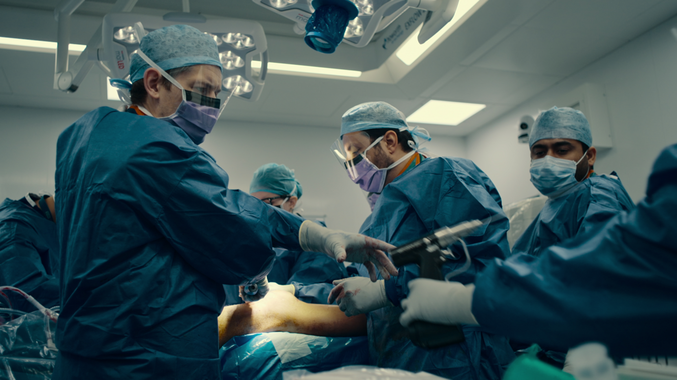 Medical staff in blue scrubs are performing a surgery in a white room. There is a person on an operating table and people passing equipment to each other.