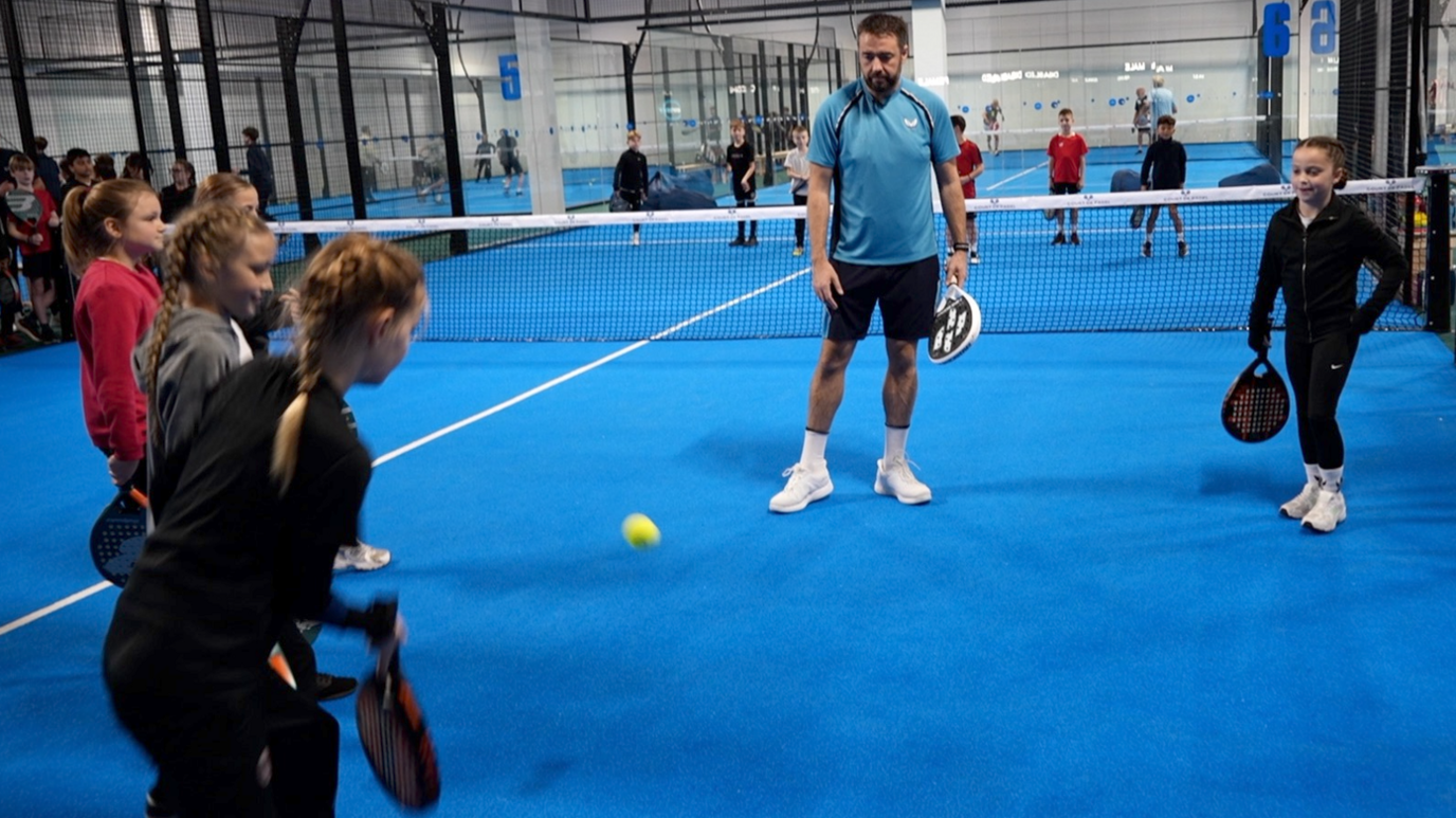 Jason Manford standing on a blue indoor tennis court watching a girl aged about 10 hit a tennis ball towards him with a padel racket. He has brown short hair and a brown beard. He is wearing a sky blue T-shirt, the same colour as the court, with black stripes down the sides, black shorts and white trainers. The girl is dressed all in black, with four other girls standing on the court, as if waiting their turn.