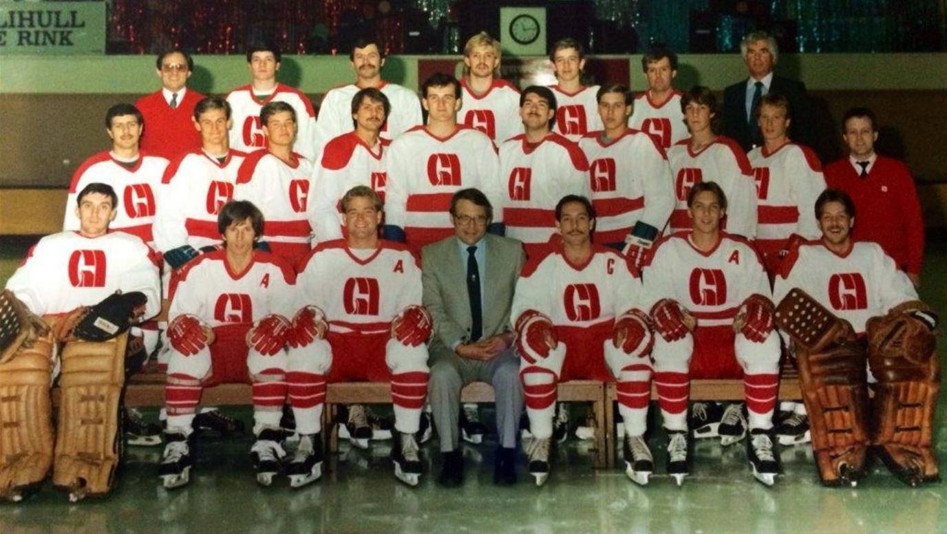 An ice hockey team photo with the player wearing large white hockey jerseys with red logos and lettering and a man in a grey suit with glasses sat in the centre