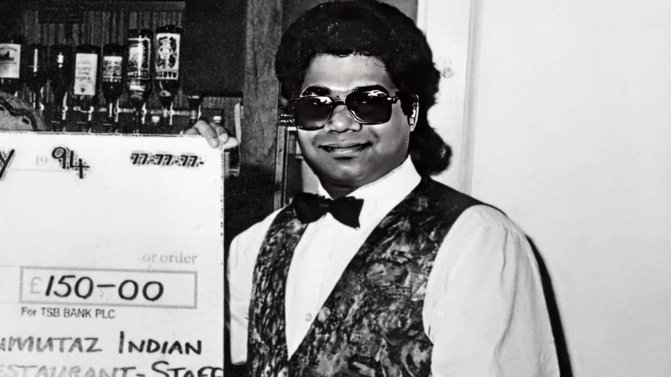 Black and white photograph of a man in a restaurant holding a big cheque. He is smiling. The man has long, dark, swept back hair. He is wearing a white shirt, black bow tie and a patterned waistcoat.