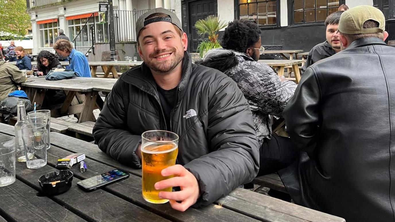 Lewis Symthe sitting at a bench and smiling, holding a pint of beer