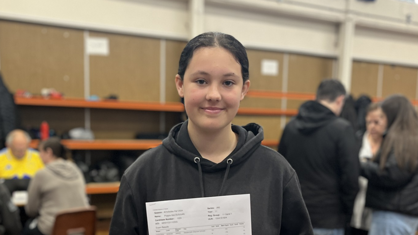 A young girl smiling and holding a piece of paper to the camera. She is wearing a black hoodie with her hair tied back