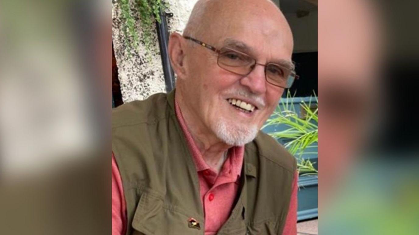 Stuart McPherson is wearing a red shirt and green jacket. He is smiling at the camera and is largely bald and has a short grey beard