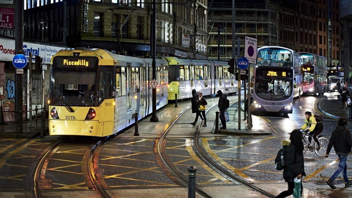 A Metrolink at nighttime travelling near Shudehill. Commuters are in the picture, some on foot and one cyclist is also in shot. Buses can be seen in the background.