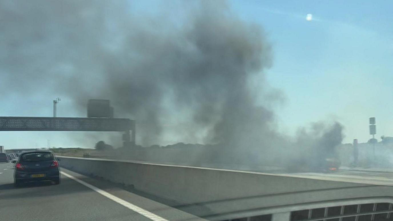 A photo taken from inside a car from a passenger seat. Dark smoke billows from a vehicle parked on the opposite carriageway, which has no other cars on. There is blue skies.