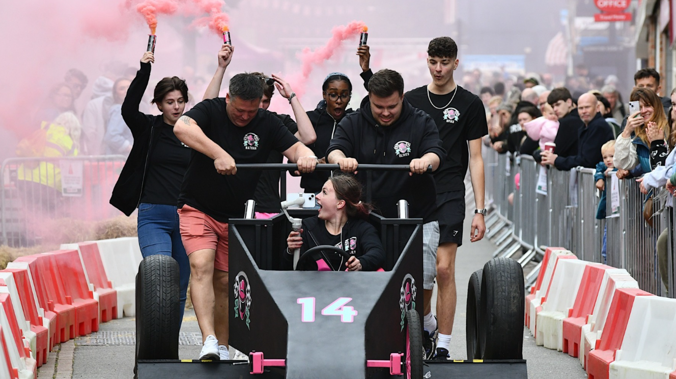 A group of people, some with flares in the background, on a race track pushing a black soapbox car with pink flashes