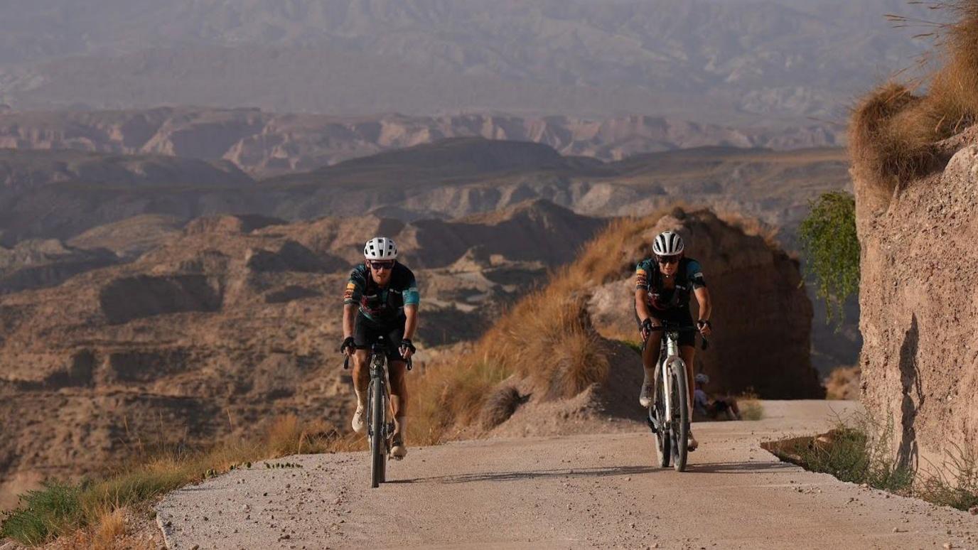 A man and a woman wearing matching cycling gear, cycling next to each other on a steep road on a sunny day with a mountainous landscape behind them.