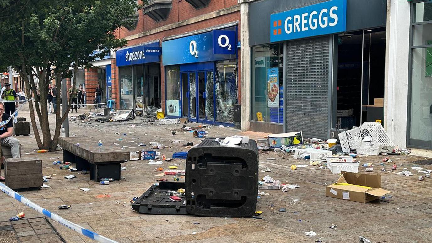 Rubble strewn across the ground in front of damaged shops, some with their windows smashed