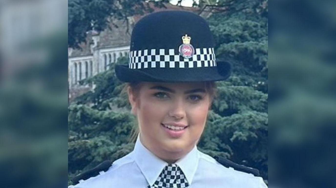 Hannah Byrne looks at the camera in her policing uniform. She is wearing a hat with the Surrey Police logo on it and a black and white checked neckerchief.